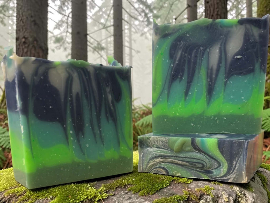 Two handmade marbled soap bars with green and dark navy swirls, displayed on a mossy log in a misty forest setting.
