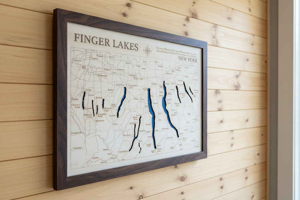 A framed wooden map of Finger Lakes, New York, mounted on a light gray wall. The map features natural wood tones with dark blue laser-etched lake channels and a dark brown wood frame.
