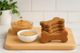 Gluten-Free Peanut Butter Bones, handmade organic dog treats from The Chosen Spot K9 Kitchen on a wooden board with a bowl of brown rice and a container of peanut butter.
