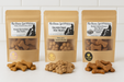 Three packages of 'The Chosen Spot K9 Kitchen' dog treats, Peanut Butter Bones, Salmon Paws and Bacon Cheddar Biscuits with Duvall Farms Bacon on a beige background. Handmade organic dog treats.