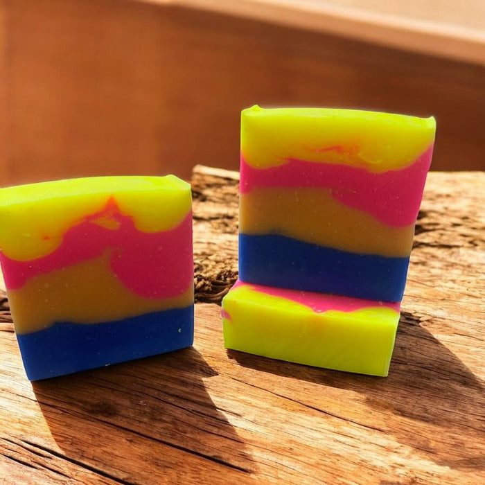 Two bars of soap with a layered pattern of yellow, pink, and blue colors, sitting on a wooden surface.