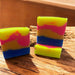 Two bars of soap with a layered pattern of yellow, pink, and blue colors, sitting on a wooden surface.